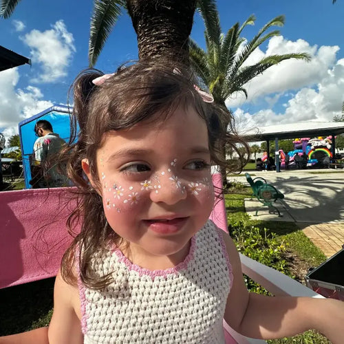 Little girl showing off a daisy flower face painting design at a sunny park event in Miami.