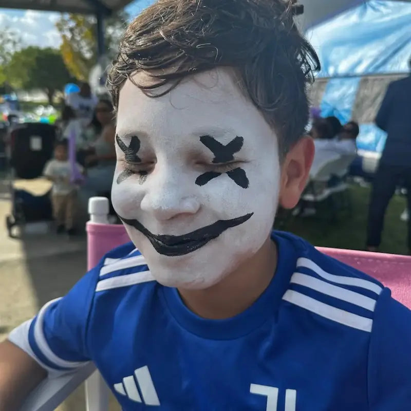 Boy showing a spooky Halloween face painting design with white base and black stitched smile at a Miami outdoor event.