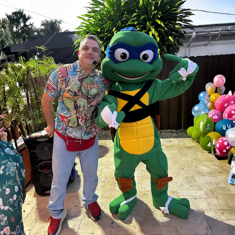 Party host with headset mic and a Ninja Turtle mascot flexing together at a TMNT themed backyard party in Miami, animation plus character combo.