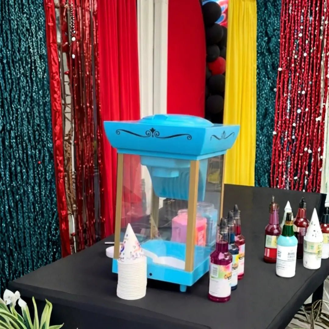Shaved ice station with blue snow cone machine, syrup flavors and paper cones in front of a colorful fringe backdrop in Miami.