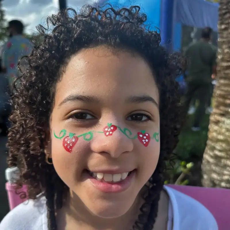 Smiling girl with a strawberry vine face painting design across her cheeks at a sunny outdoor event in Miami.