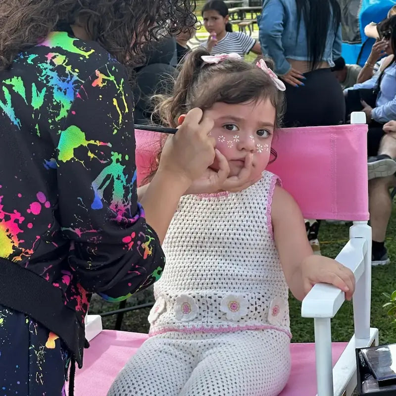 Close-up of a toddler getting daisy face paint applied with a fine brush at an outdoor family event in Miami.